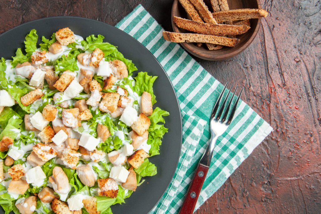A fresh Caesar Salad in a dark bowl featuring bright green romaine leaves, golden sourdough croutons, and large shards of Parmesan cheese under soft lighting.